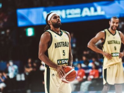 An Indigenous basketball player wearing an Australian uniform holds a basketball and prepares to shoot. There is a teammate and a stadium crowd in the background.